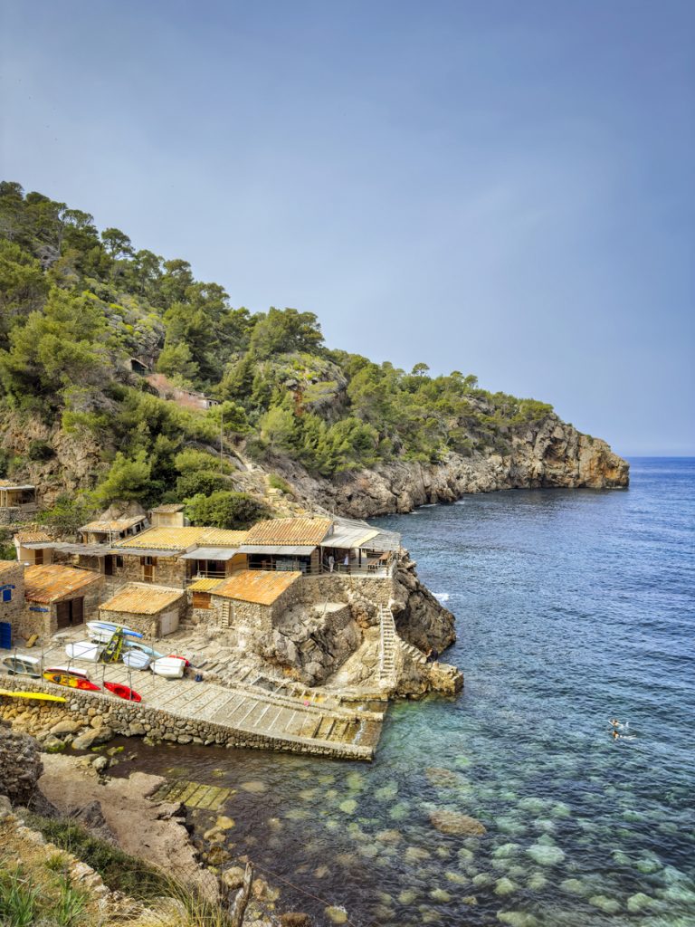 Steinige Badebucht Cala Deià mit klarer türkisblauer Wasserfläche und Bootsgaragen an der Küste Mallorcas