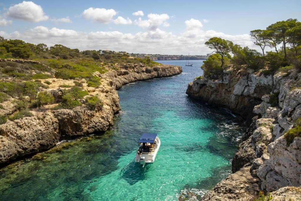 Boot in der türkisfarbenen Bucht Cala Beltrán bei Llucmajor auf Mallorca, umgeben von Felsen und Pinien.