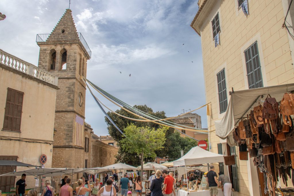 Blick auf den belebten Wochenmarkt in Son Servera auf Mallorca mit Marktständen, Lederwaren und dem historischen Kirchturm im Hintergrund.