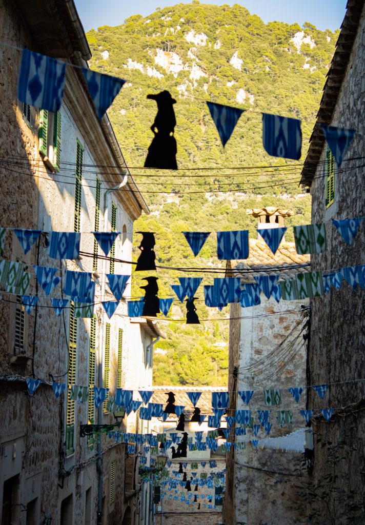 Schmale, mit blau-weißen Wimpeln und schwarzen Silhouetten geschmückte Gasse in Valldemossa, Mallorca, mit Blick auf die umliegenden grünen Berge.