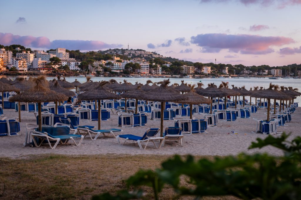 Blick auf den Strand von Santa Ponsa auf Mallorca mit leeren Sonnenliegen und Strohdach-Sonnenschirmen, im Hintergrund das ruhige Meer und Küstengebäude bei Abendlicht.
