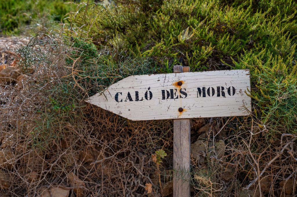 Holzschild mit der Aufschrift „Caló des Moro“ vor dichter, trockener Vegetation