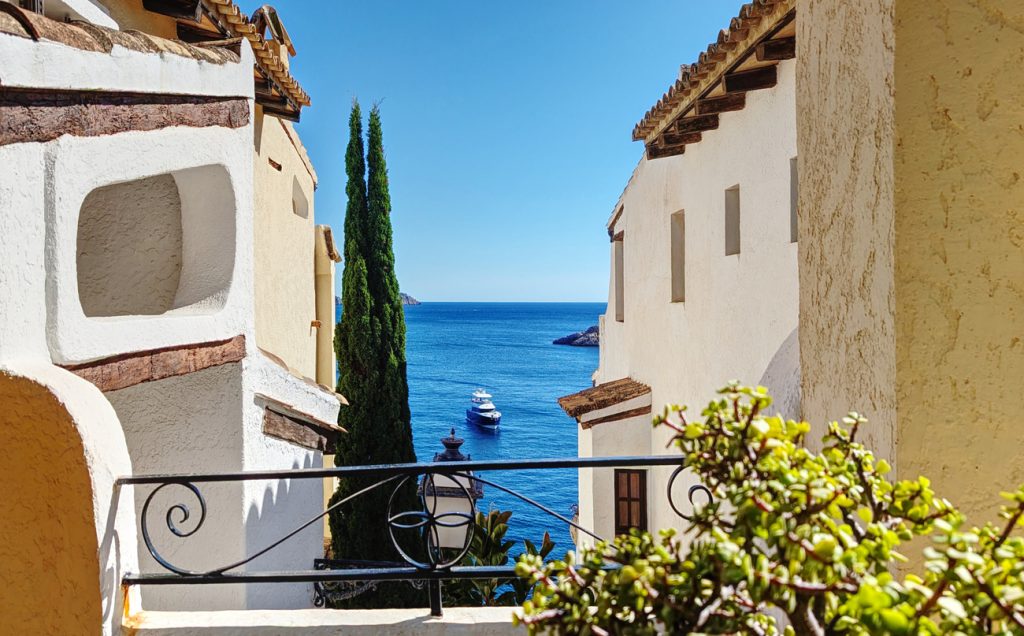 Blick durch eine Gasse in Cala Fornells auf das tiefblaue Meer mit einer Yacht, eingerahmt von mediterranen Häusern und Zypressen