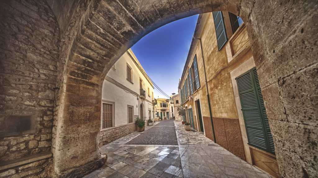 Steinbogen mit Blick auf eine malerische Gasse in der Altstadt von Santanyí auf Mallorca mit traditionellen Sandsteinhäusern und grünen Fensterläden.