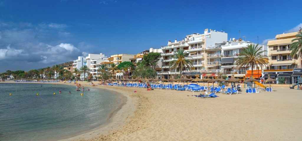 Strand von Port de Pollença mit Sonnenschirmen, Liegen und Blick auf die Uferpromenade mit Palmen und Wohnhäusern