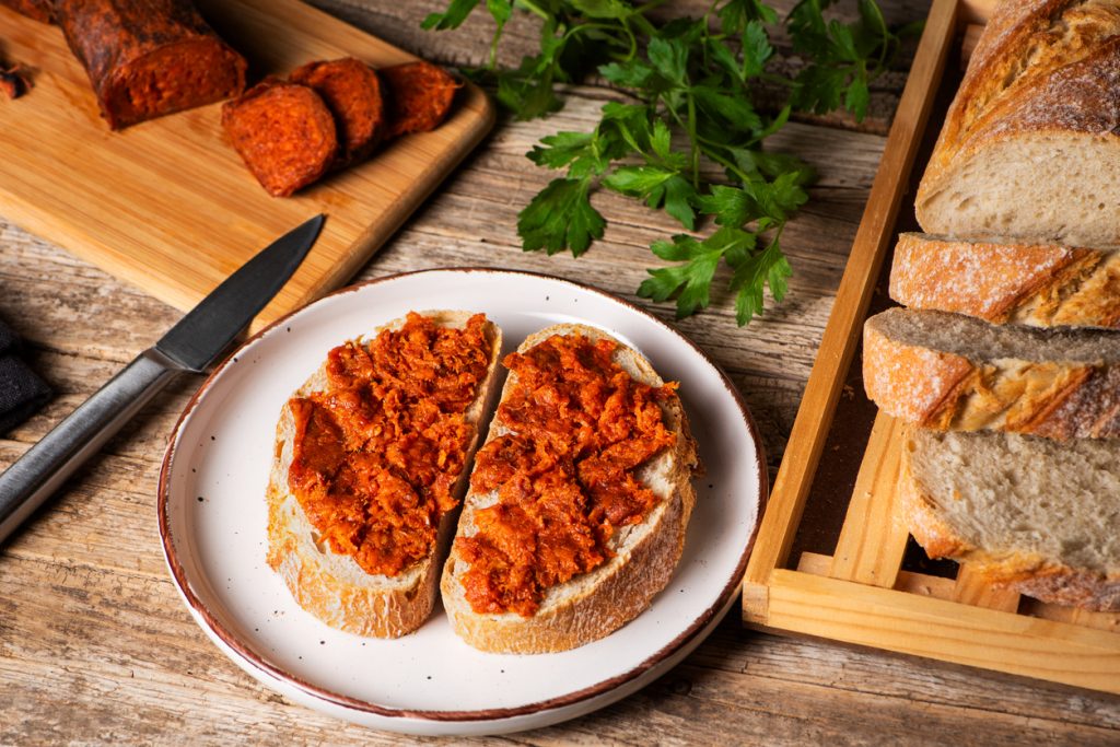 Zwei Scheiben Brot mit Sobrasada-Aufstrich auf einem Teller, daneben frisches Baguette und geschnittene Sobrasada-Wurst.