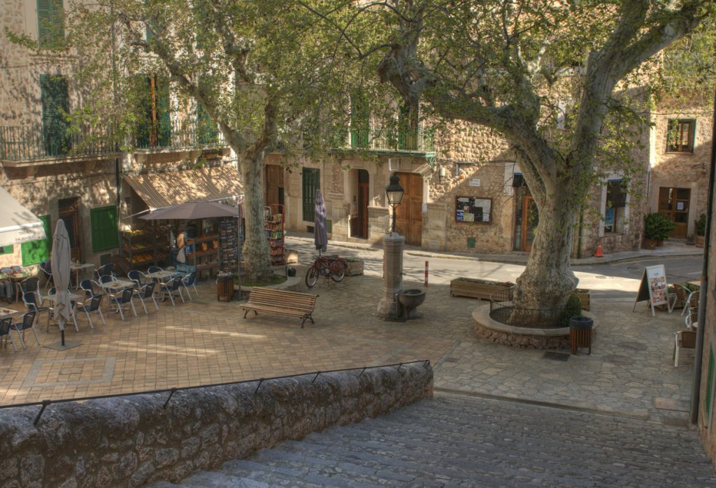 Malerischer Dorfplatz in Fornalutx auf Mallorca mit Caféstühlen, großen Platanen und traditionellen Steingebäuden.