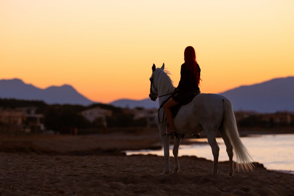 Frau mit roten Haaren reitet auf einem weißen Pferd am Strand von Mallorca in der Abenddämmerung