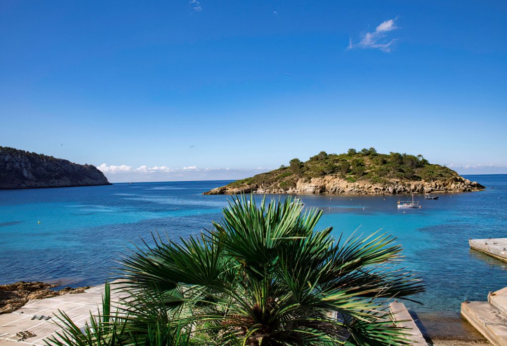 Vorgelagerte Insel Illa Es Pantaleu vor Sant Elm auf Mallorca, umgeben von türkisblauem Wasser, mit Booten und grüner Vegetation, im Hintergrund der Weg zur Insel Dragonera.