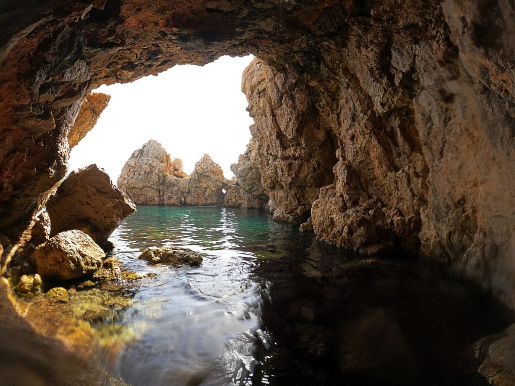 Blick aus einer Felsgrotte auf das klare türkisfarbene Meer und die steilen Felsen der Insel Dragonera bei Mallorca.
