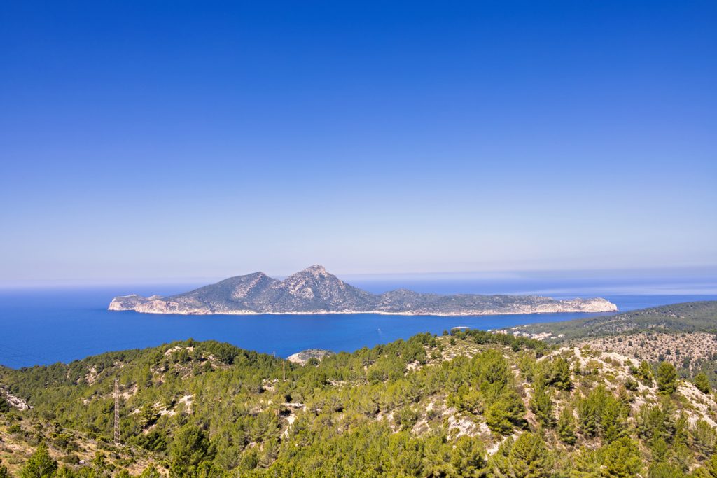 Weitwinkelaufnahme der Insel Dragonera vor Mallorca, umgeben vom tiefblauen Mittelmeer, im Vordergrund grüne Pinienlandschaft und sanfte Hügel.
