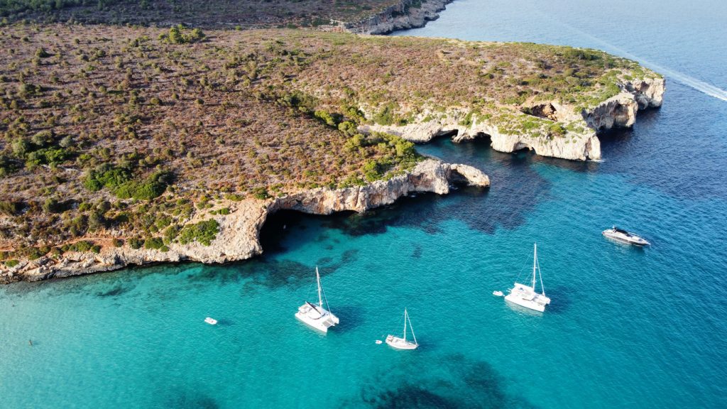 Luftaufnahme der Cala Varques auf Mallorca mit türkisblauem Wasser, Klippen und mehreren Segelbooten.