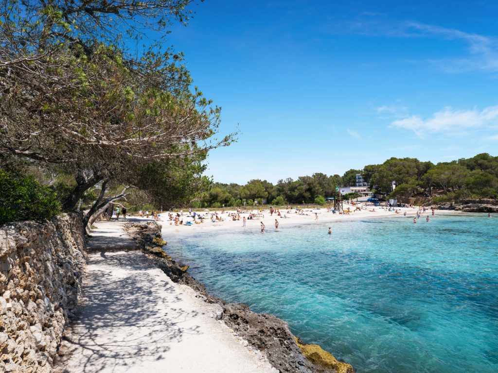 Feinsandiger Strand Cala Mondragó mit türkisblauem Wasser, Pinienwäldern und Badegästen im Naturpark Mondragó auf Mallorca.