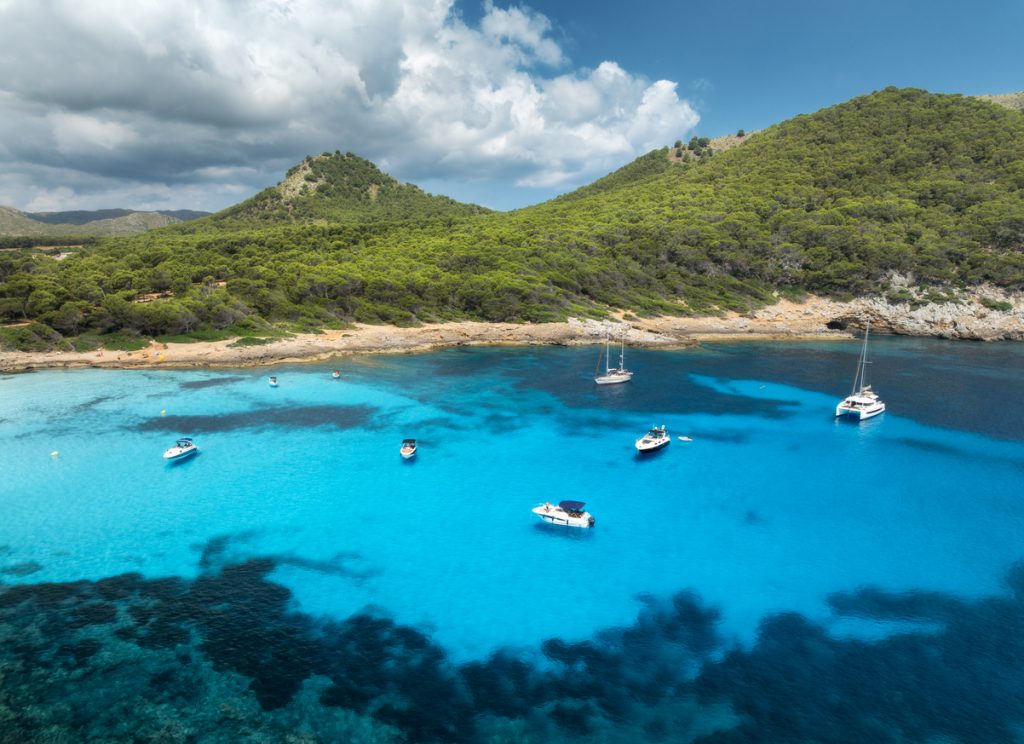 Segel- und Motorboote liegen im klaren, türkisblauen Wasser vor der Cala Agulla auf Mallorca, umgeben von grünen Hügeln und felsiger Küste.