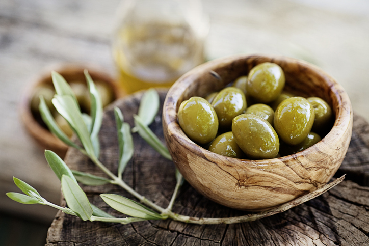 Schale aus Olivenholz mit frischen grünen Oliven auf rustikalem Holztisch, daneben ein Olivenzweig