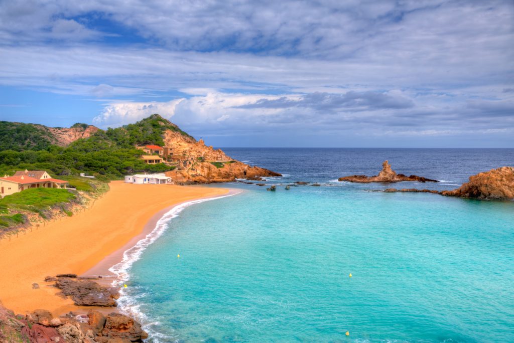 Goldener Sandstrand mit türkisfarbenem Wasser in der Bucht Cala Pregonda auf Menorca bei leicht bewölktem Himmel