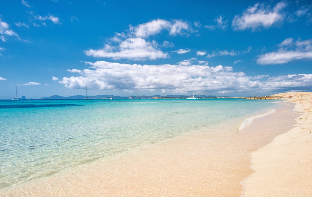 Feiner weißer Sandstrand am Playa de Ses Illetes auf Formentera mit türkisblauem Wasser und Segelbooten im Hintergrund.