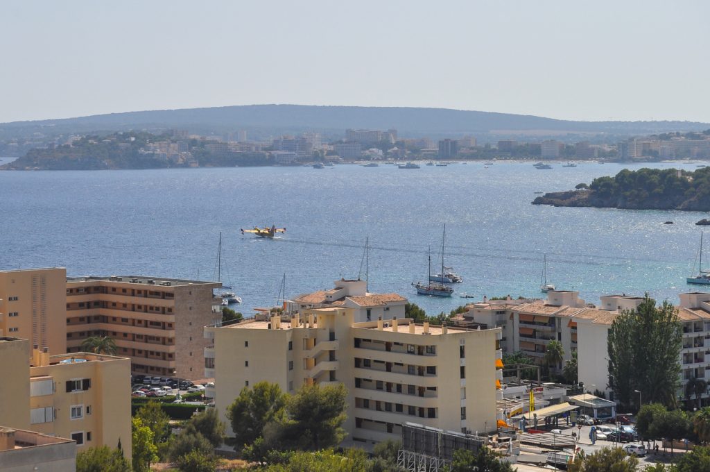 Ein gelbes Löschflugzeug nimmt Wasser aus der Bucht vor Mallorca auf, im Vordergrund Wohngebäude und Boote