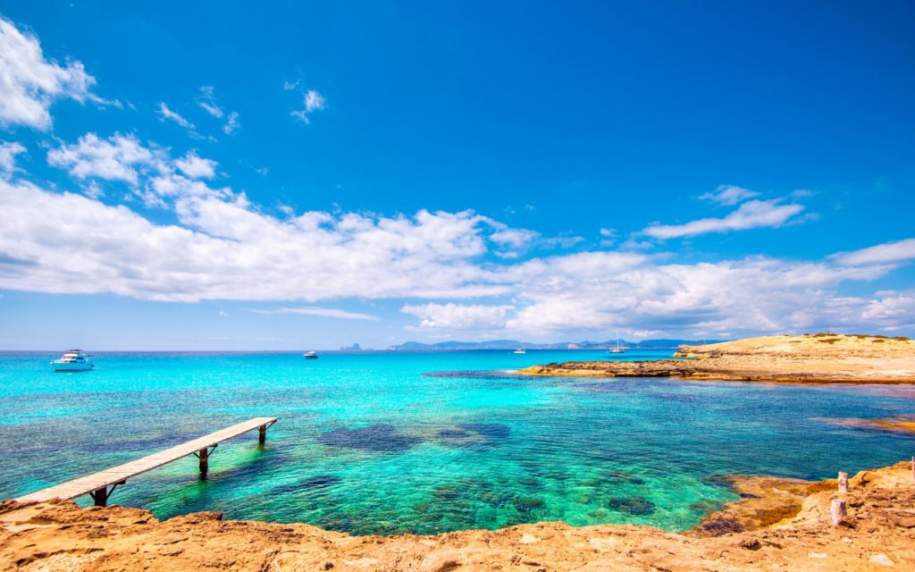 Holzsteg ragt ins türkisfarbene Meer der Playa de Ses Illetes auf Formentera, im Hintergrund Boote und die Küste unter blauem Himmel.