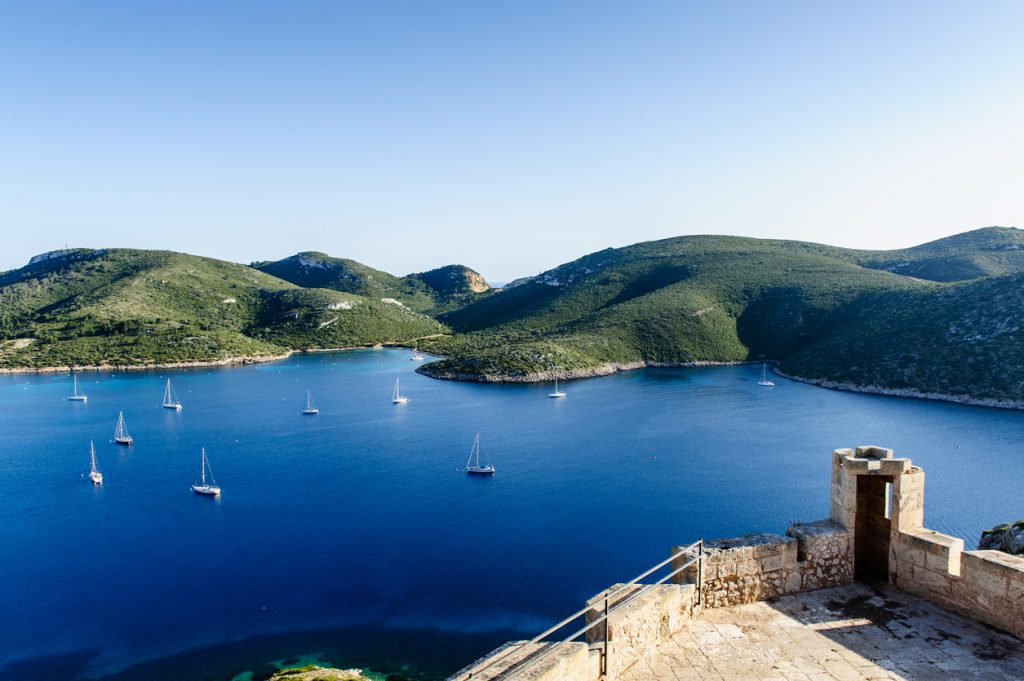Mehrere Segelboote liegen vor Anker in der geschützten Bucht von Cabrera, im Vordergrund ein Aussichtspunkt mit alten Mauern