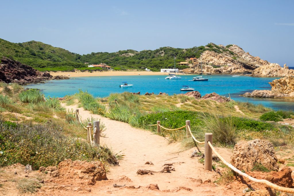 Sandweg mit Blick auf die türkisfarbene Bucht Cala Pregonda auf Menorca mit Booten im Wasser