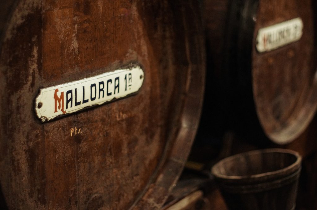 Nahaufnahme eines alten Holzfasses in einer Bodega auf Mallorca mit Metallschild „Mallorca 1ª“.