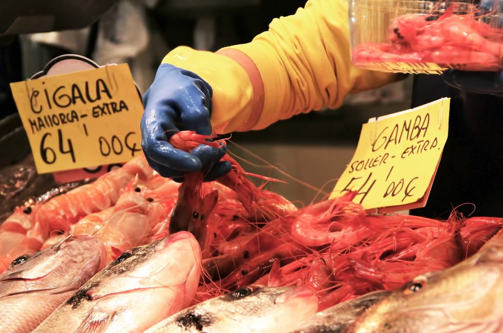 Nahaufnahme von roten Garnelen und frischem Fisch auf einem Marktstand in Sóller, Mallorca, mit Preisschildern und einer Person mit blauen Handschuhen, die Garnelen sortiert.