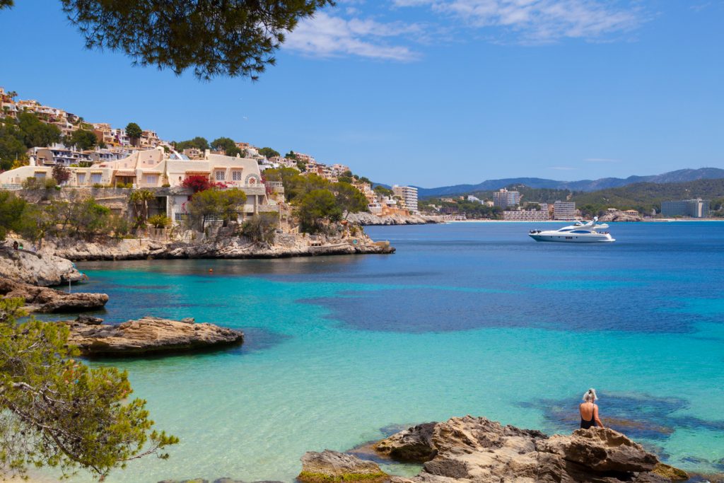 Blick auf Cala Fornells bei Peguera auf Mallorca mit türkisfarbenem Meer, Felsen, Häusern und einer Yacht im Hintergrund.