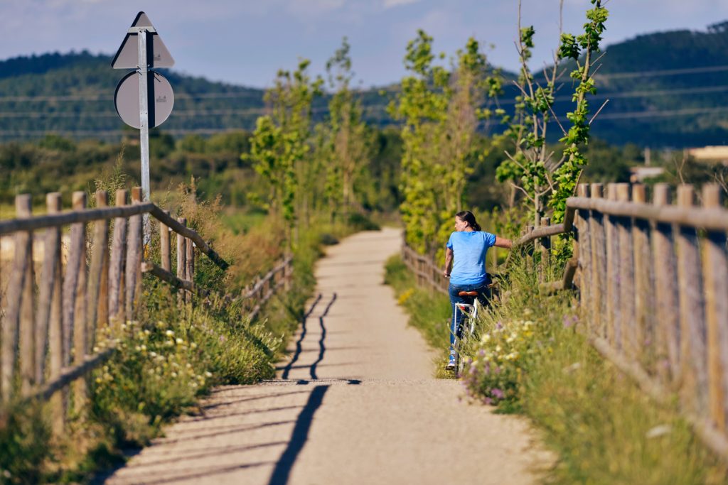 Frau fĂ€hrt mit dem Fahrrad auf der Via Verde, einem landschaftlich schönen Radweg mit HolzzĂ€unen und grĂŒner Umgebung.