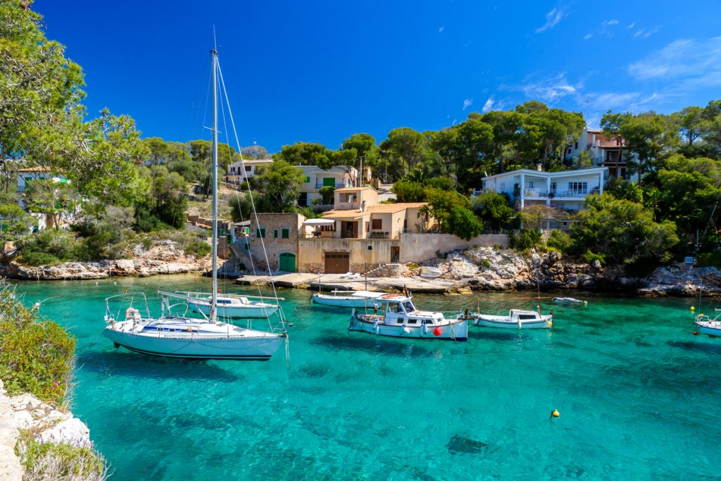 Traditionelle Boote und Segelyachten liegen im kristallklaren Wasser der Cala Figuera auf Mallorca, umgeben von Häusern und Pinienwäldern.