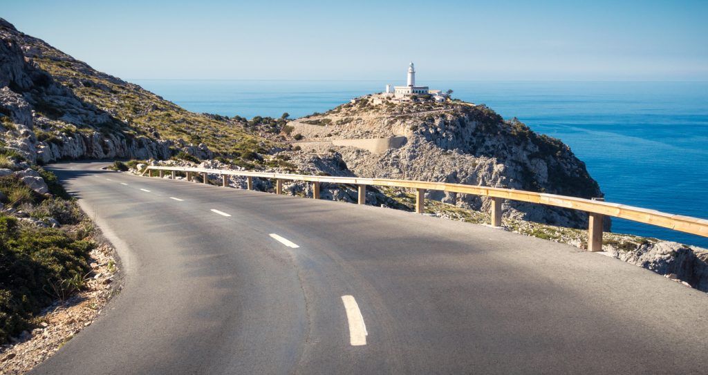Blick auf die kurvenreiche Küstenstraße zum Leuchtturm am Cap Formentor auf Mallorca, mit Meer und Felslandschaft im Hintergrund.
