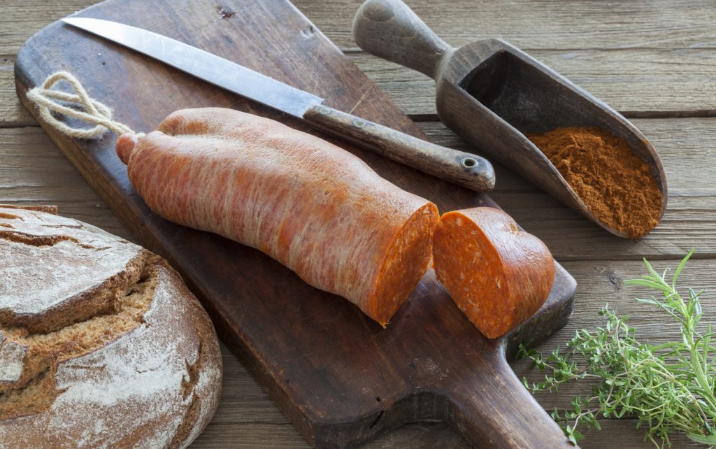 Traditionelle Sobrasada-Wurst aus Mallorca auf einem Holzbrett mit Brot, Messer, Paprikapulver und frischen Kräutern.