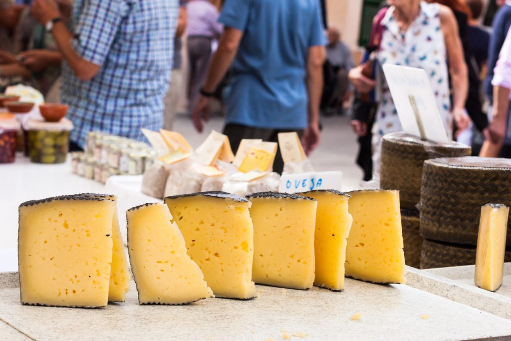 Frische Stücke Manchego Käse auf dem Wochenmarkt in Sineu auf Mallorca, im Hintergrund Marktbesucher und regionale Produkte.