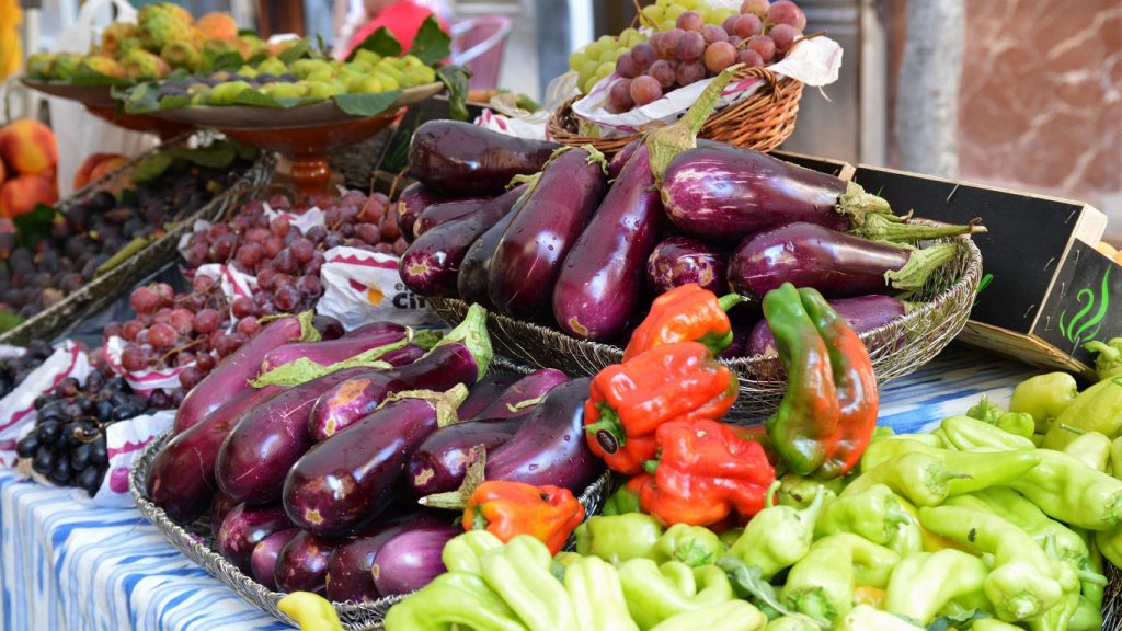 Nahaufnahme von frischem Gemüse und Obst auf einem Marktstand auf Mallorca, darunter Auberginen, Paprika, Weintrauben und Feigen, dekorativ auf Tischen mit blau-weiß gestreifter Tischdecke arrangiert.