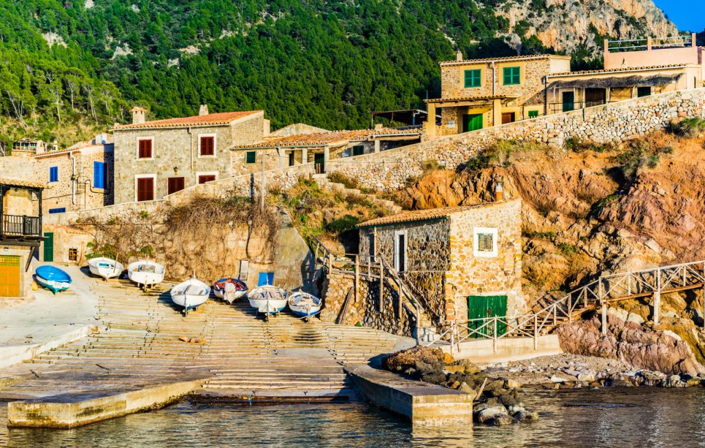 Kleine Boote und traditionelle Steinhäuser am Hafen von Valldemossa, Mallorca, mit Blick auf die umliegenden grünen Berge.