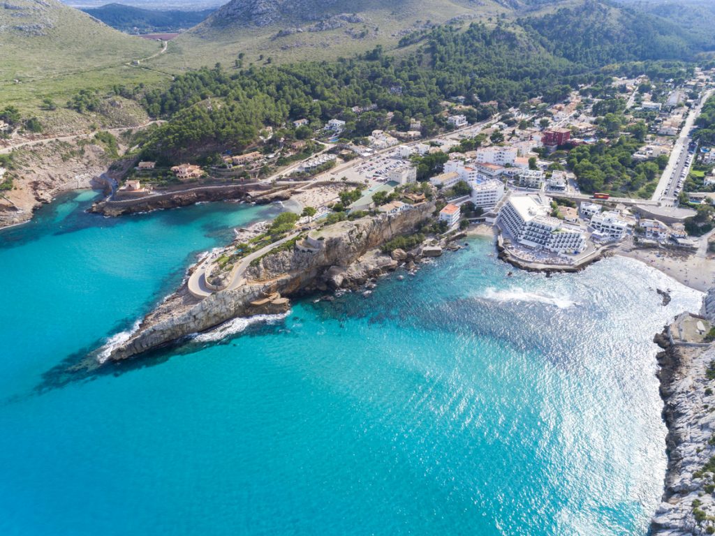 Luftaufnahme von Cala Sant Vicenç mit Strand, Hotelgebäuden, Küstenstraße und türkisfarbenem Meer