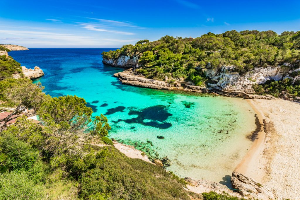 Luftaufnahme der Cala Llombards mit weißem Sandstrand, türkisem Wasser und umgebenden Pinienhügeln