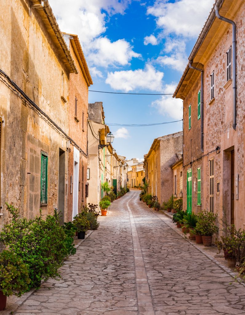 Schmale, gepflasterte Gasse in Petra auf Mallorca, gesäumt von traditionellen Sandsteinhäusern mit grünen Fensterläden und Topfpflanzen vor blauem Himmel.