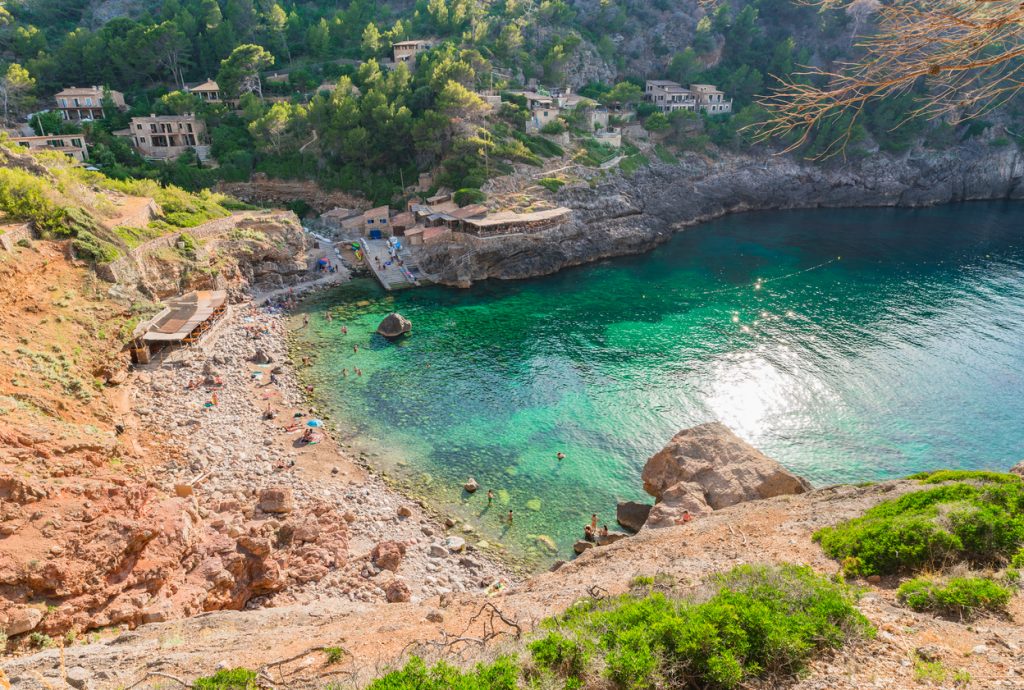 Sand- und kiesbedeckte Cala Deià mit kristallklarem, türkisblauem Wasser zwischen hohen Felswänden und Kiefern