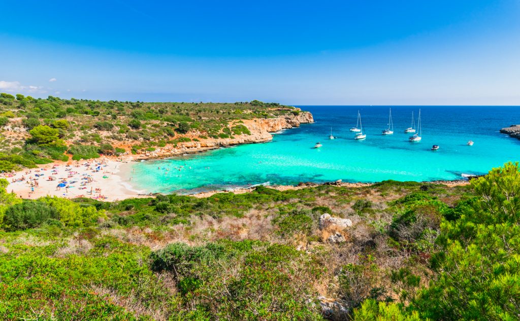 Cala Varques auf Mallorca mit weißem Sandstrand, türkisfarbenem Wasser, Badenden und mehreren Segelbooten im Meer.