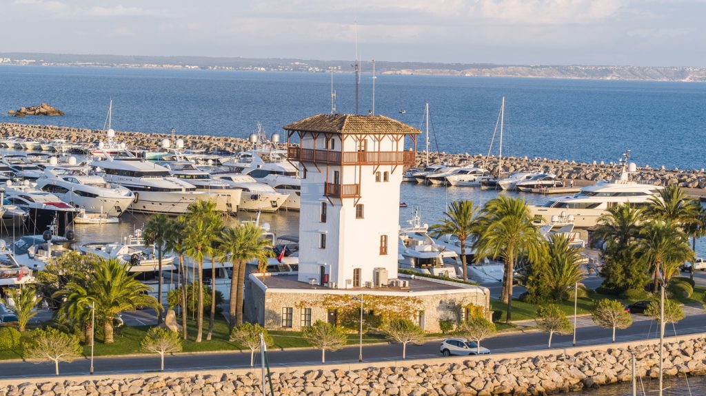 Historischer Turm im Yachthafen Puerto Portals auf Mallorca, umgeben von luxuriösen Yachten und Palmen.