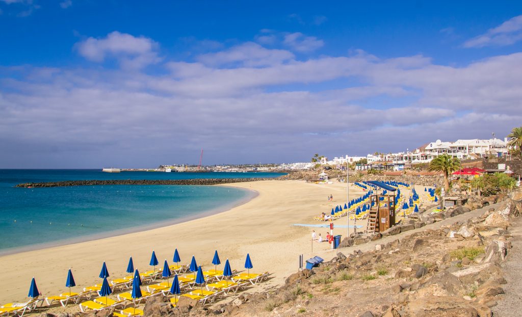 Breiter Sandstrand Playa Blanca auf Lanzarote mit gelben Liegen, blauen Sonnenschirmen und ruhigem türkisfarbenem Meer.