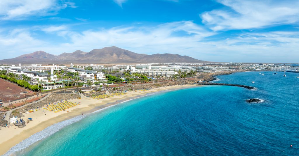 Panoramablick auf Playa Blanca auf Lanzarote mit weißen Hotels, Sandstrand, türkisblauem Meer und Bergen im Hintergrund.