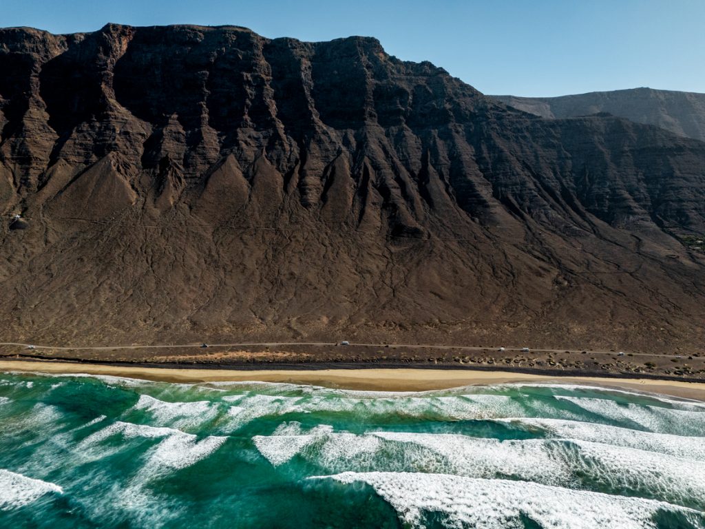Drohnenaufnahme der Playa de Famara auf Lanzarote mit türkisfarbenem Wasser, Wellen und den steilen Klippen im Hintergrund.