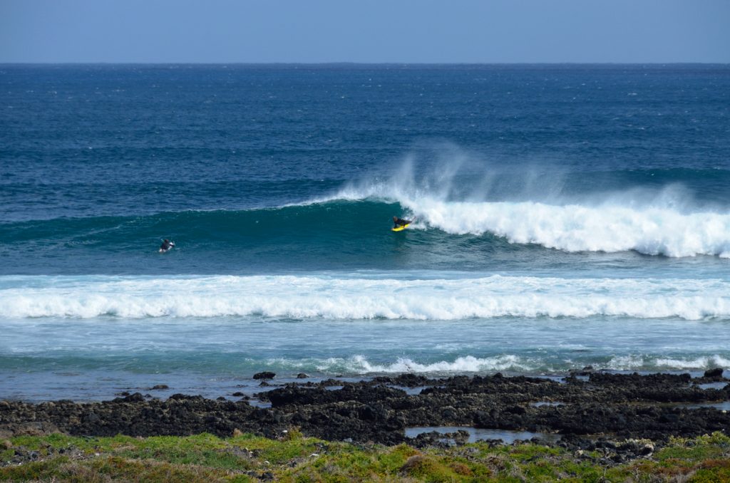 Surfer am Caletón Blanco auf Lanzarote reitet eine Welle, im Vordergrund schwarze Lavafelsen und grüne Küstenvegetation.
