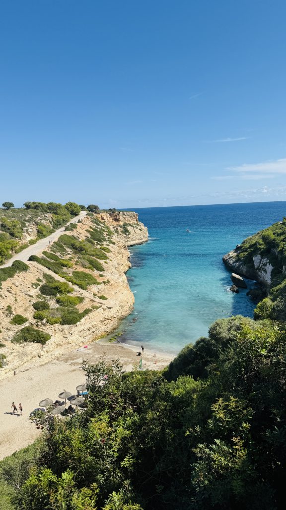 Blick von oben auf die Cala Antena: schmale Felsbucht mit klarem Türkis, kleinem Strand, wenigen Badegästen und Küstenweg auf der linken Klippe.