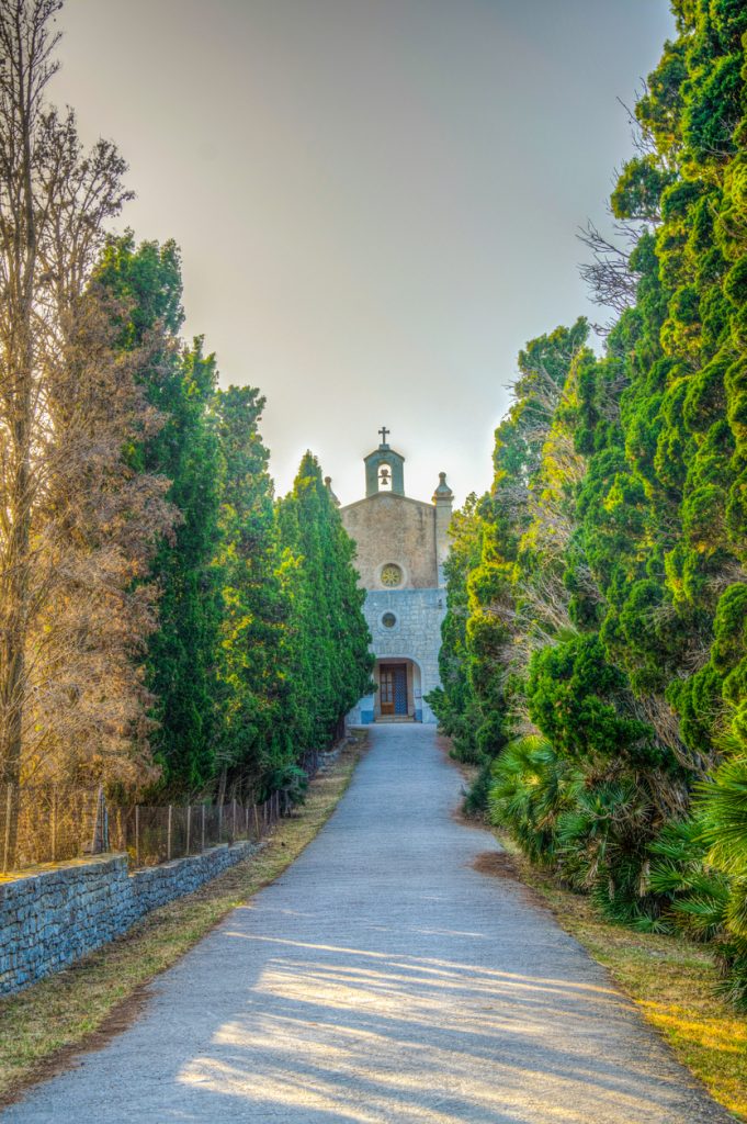Gerader Weg zwischen Zypressen zur Steinkapelle Ermita de Betlem mit Glockengiebel.