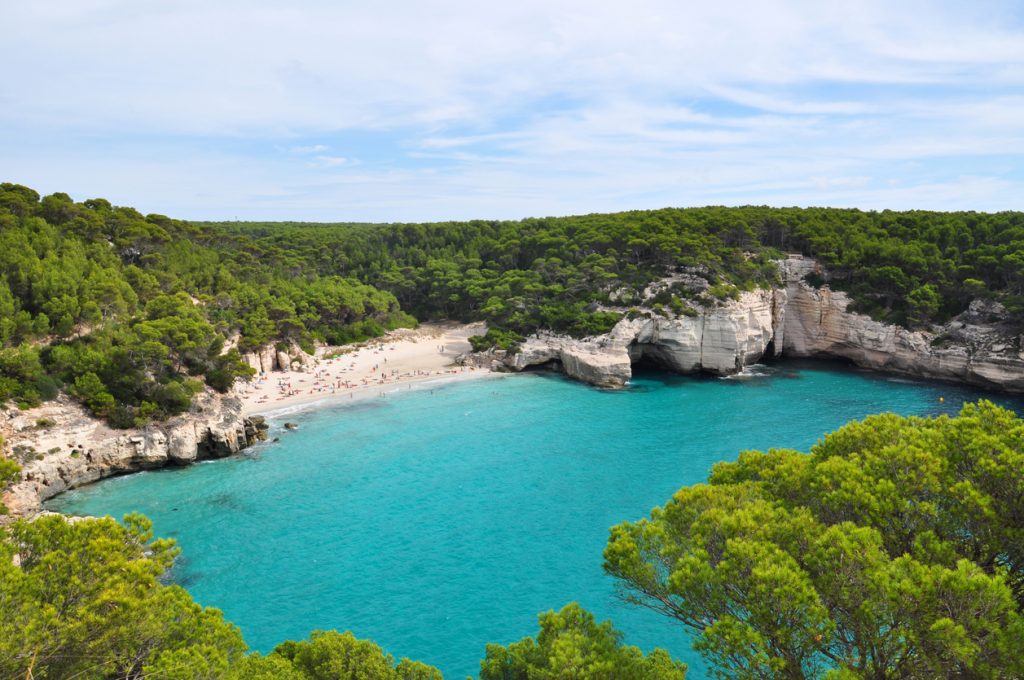 Panoramablick auf die Cala Mitjana mit weißem Sandstrand, türkisblauem Wasser, Pinienwald und markanten Kalkfelsen mit Grotte.