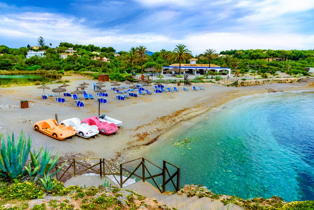 Blick auf die Cala Murada mit Strohschirmen, blauen Liegen, Tretbooten am Strand und einem Strandlokal zwischen Palmen.
