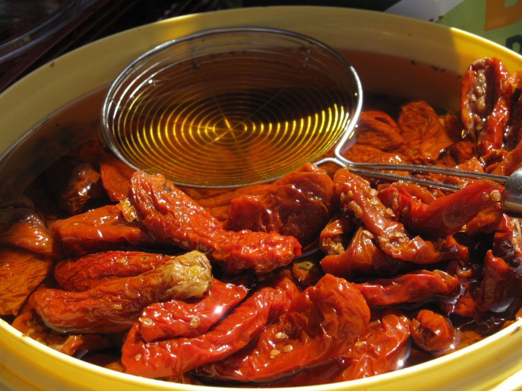 Gelbe Schüssel mit getrockneten, in Olivenöl eingelegten Tomaten sowie Metallschaumlöffel und Zange auf einem mallorquinischen Markt.